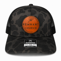 A Richardson 112 trucker hat featuring a dark gray and black camouflage pattern on the front panels and visor, black mesh back panels, and a prominent orange circular patch on the front with the text 'The REMNANT CHURCH'.