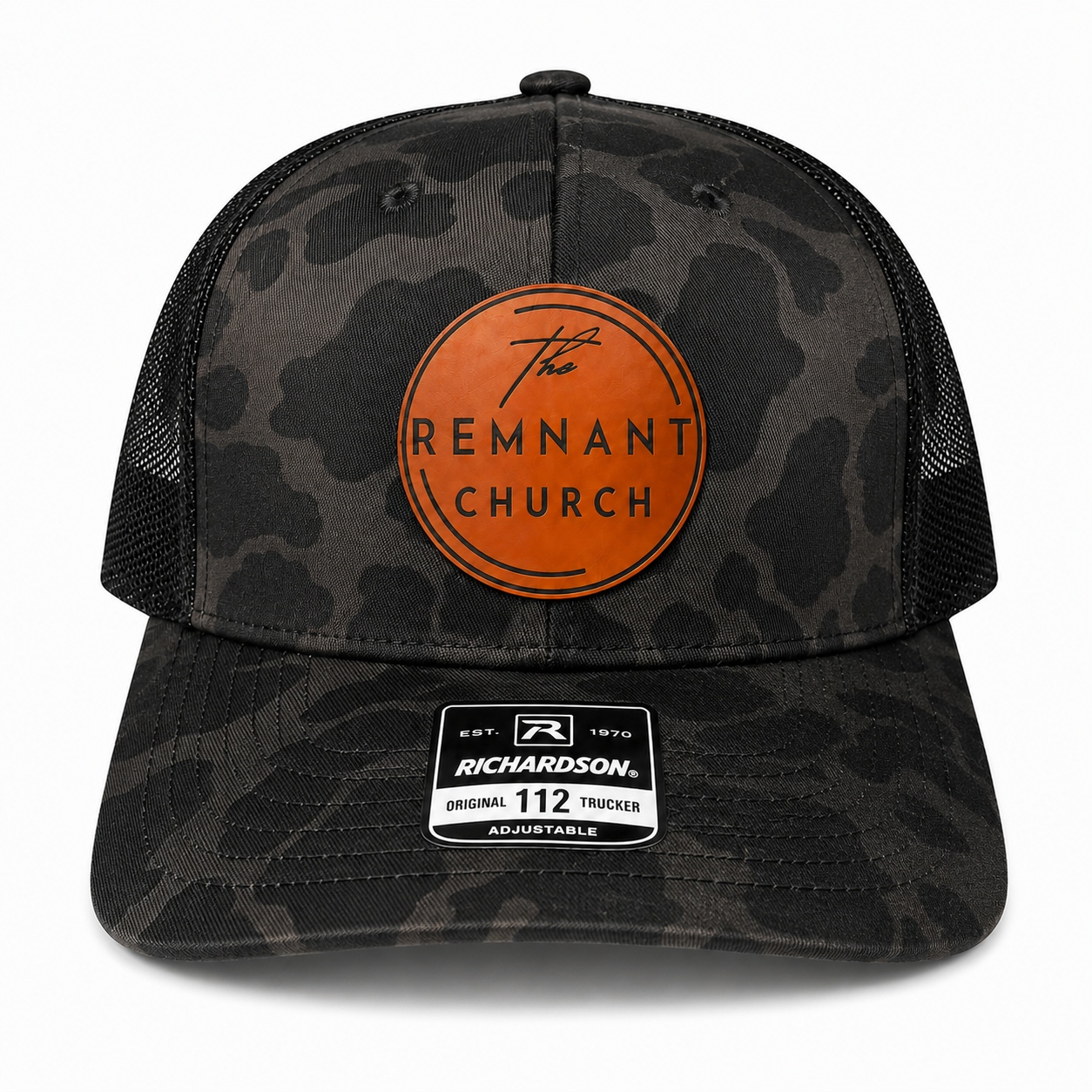 A Richardson 112 trucker hat featuring a dark gray and black camouflage pattern on the front panels and visor, black mesh back panels, and a prominent orange circular patch on the front with the text 'The REMNANT CHURCH'.