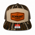 A camouflage trucker hat with a beige front panel and a prominent orange shield-shaped patch reading 'The REMNANT CHURCH.' The brim and back mesh are in a brown, beige, and black camo pattern.